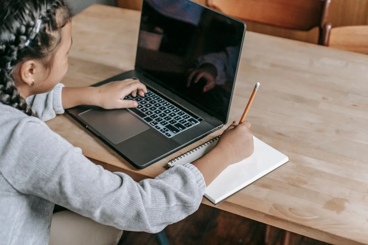 Estudiante usa una computadora portátil y toma notas en un cuaderno sobre una mesa de madera.