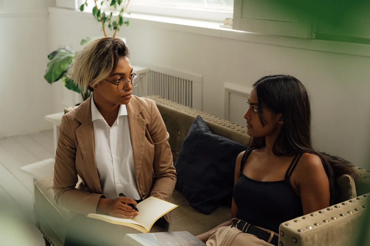 Dos mujeres conversan sentadas; una escucha mientras sostiene un cuaderno para tomar notas.