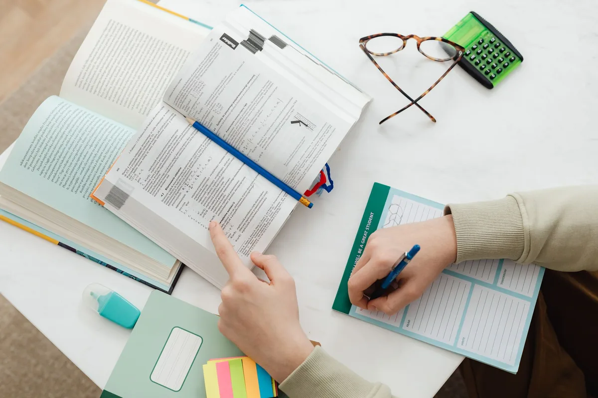 Persona repasando ejercicios de un libro y tomando notas, junto a una calculadora y cuadernos.