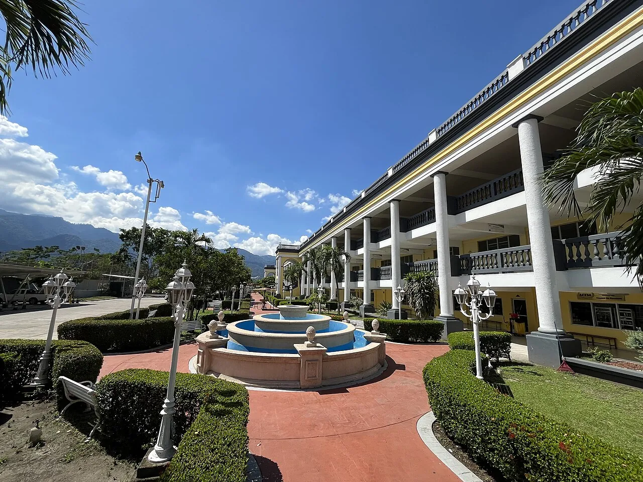 Patio del campus de UTH en San Pedro Sula, con fuente circular, jardines y un edificio amarillo de columnas blancas.
